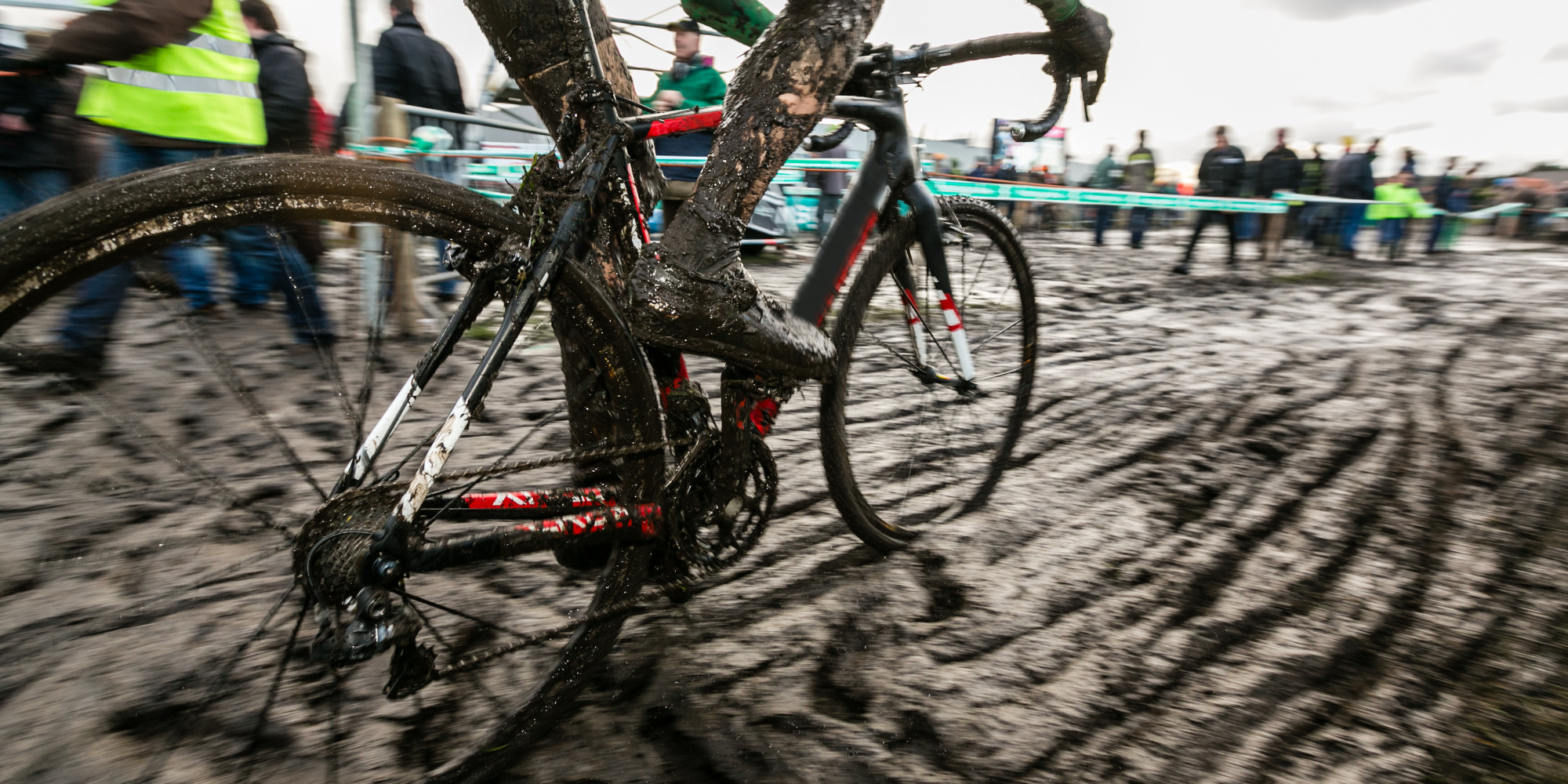 Ein Fahrrad mit schlammigen Reifen auf einem matschigen Boden, im Hintergrund Zuschauer.