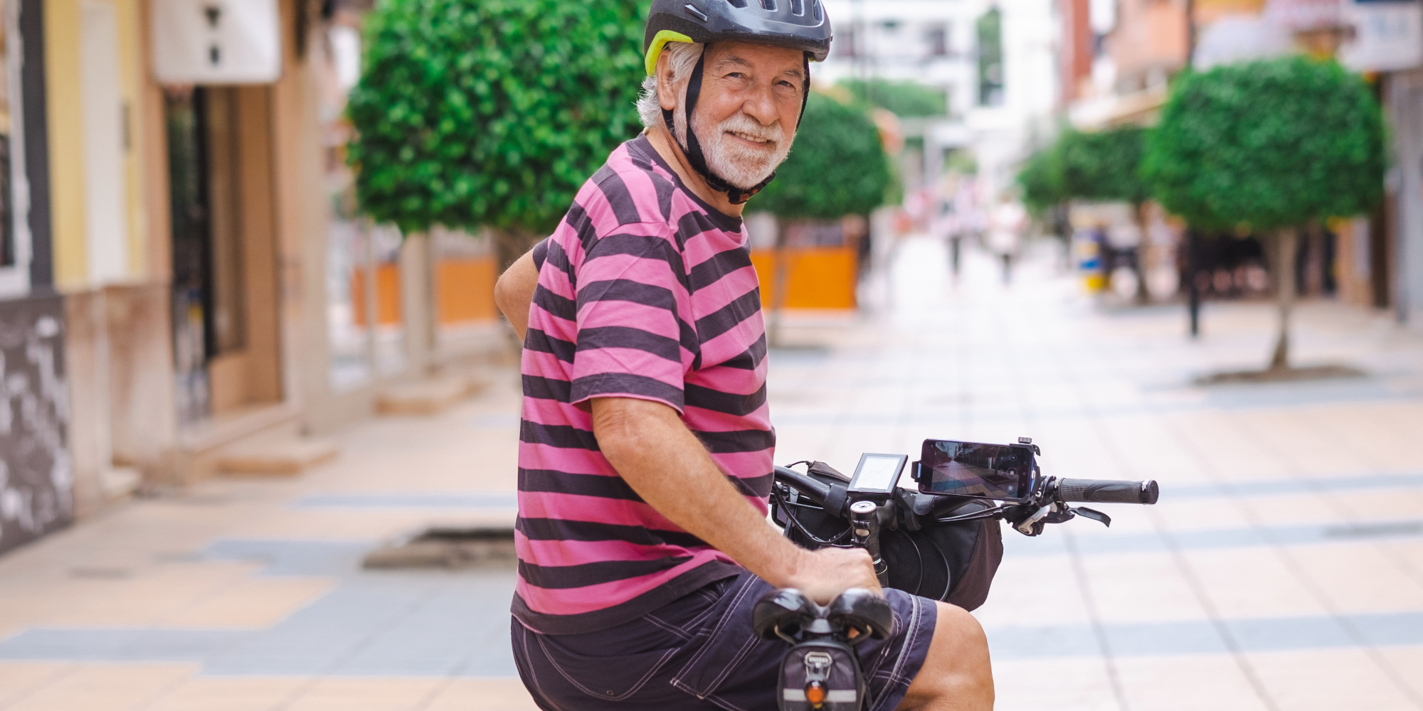 Älterer Mann in gestreiftem Shirt und Helm, lächelnd auf einem Fahrrad in einer ruhigen Straße.