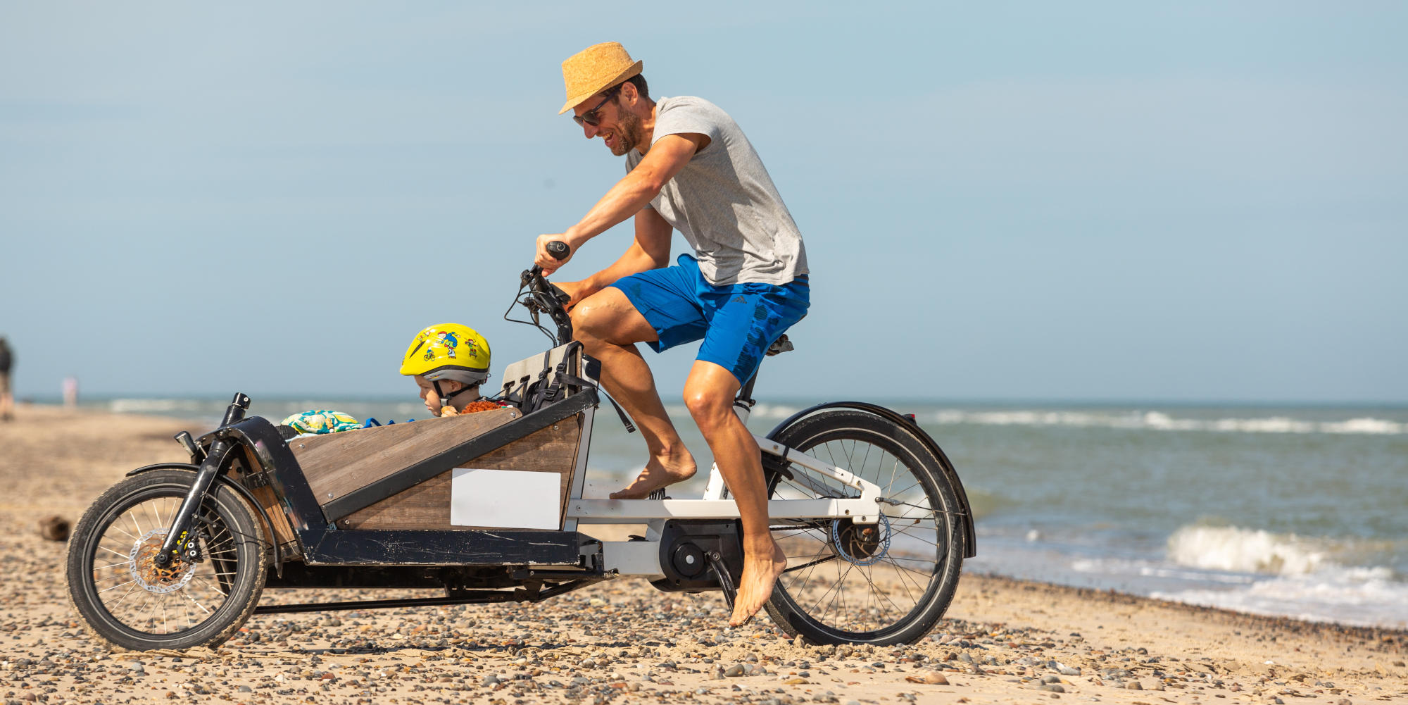 E-Lastenrad: Vorteile & Trends. Mann mit Kind und Lastenrad am Strand.