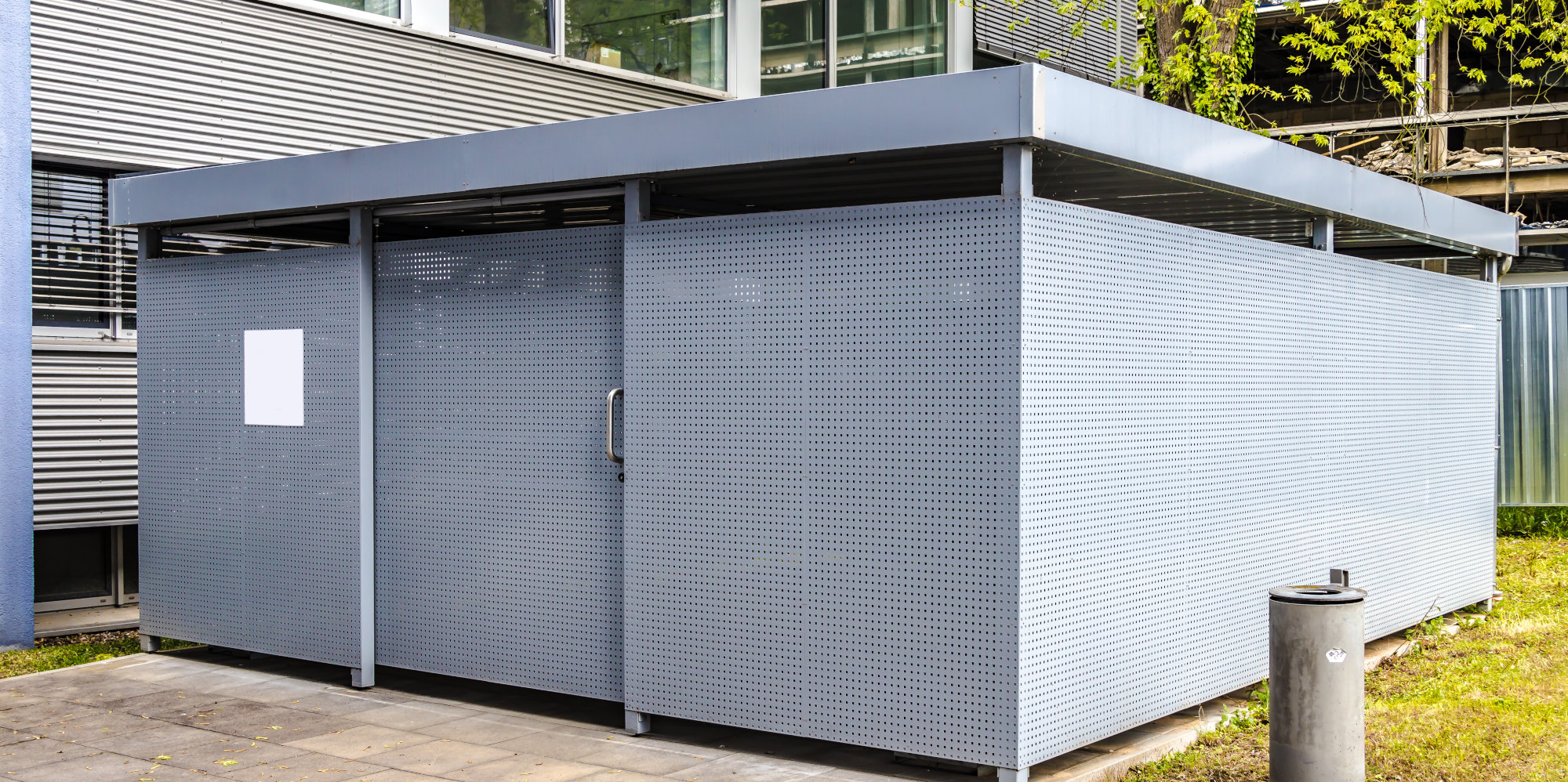 Graues, modernes Gebäude mit flachem Dach und Eingangstür, umgeben von Grünflächen.