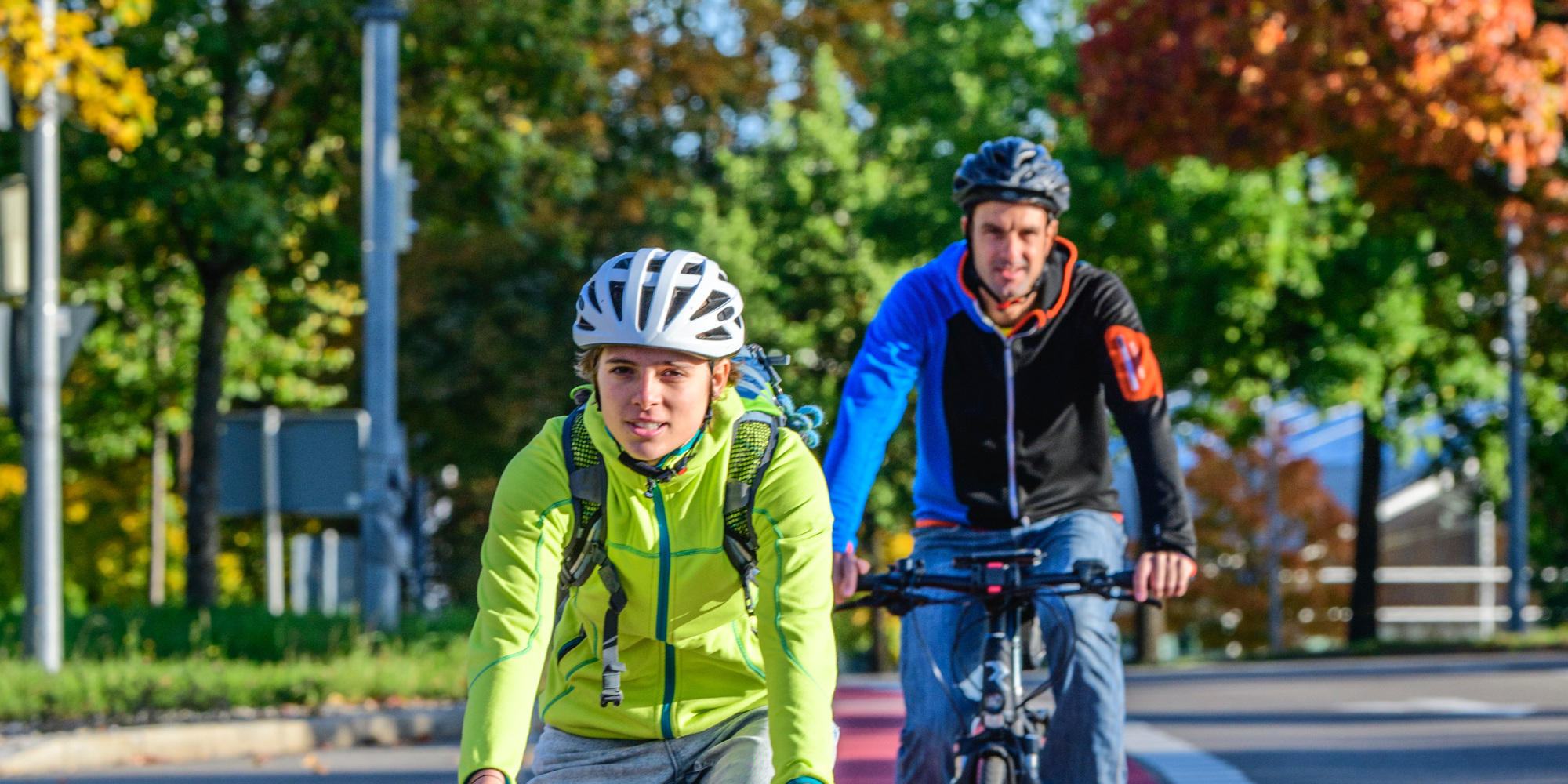 Zwei Radfahrer fahren auf einer Straße mit bunten herbstlichen Bäumen im Hintergrund.