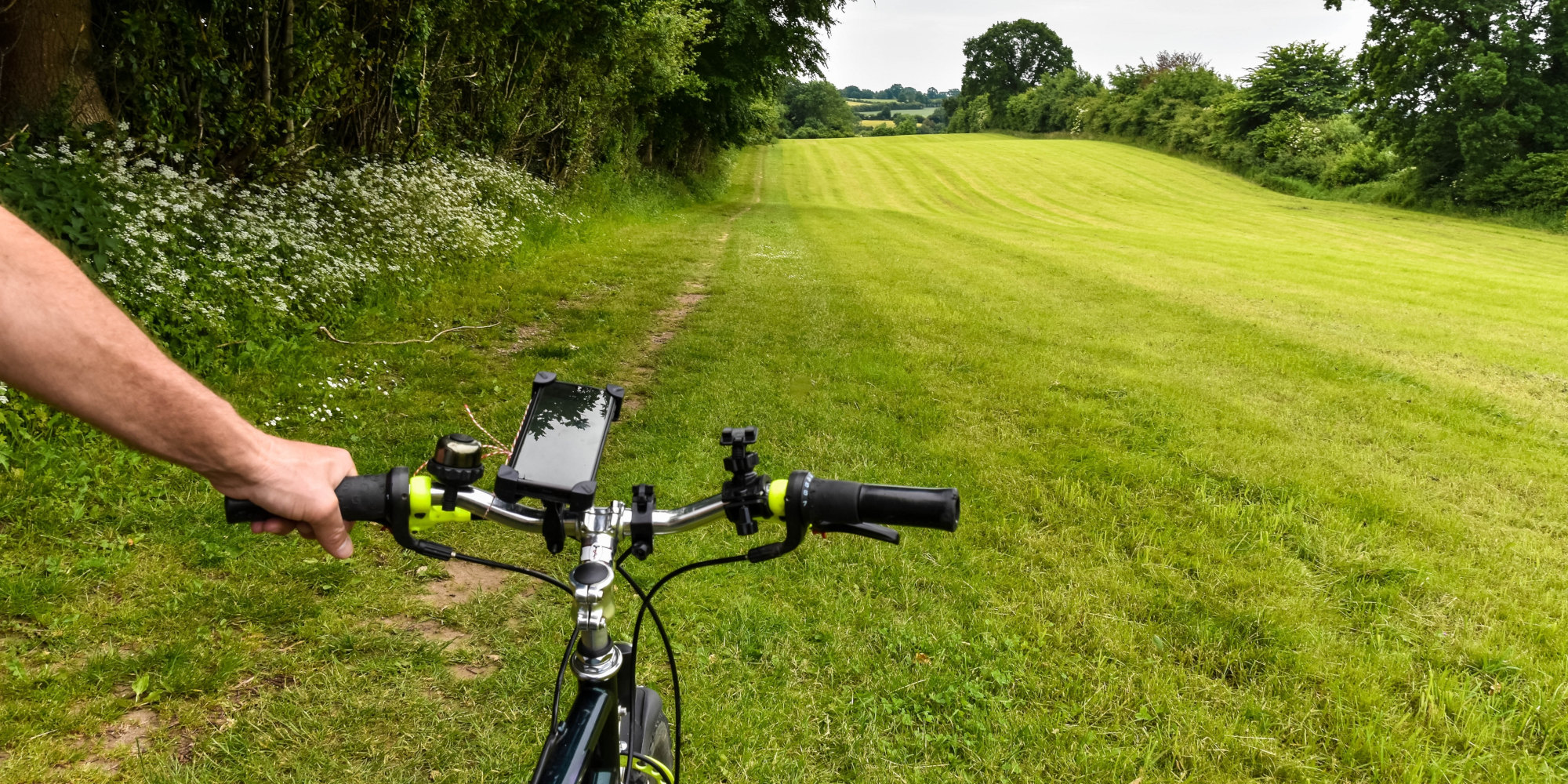 Ein Fahrradlenker mit einer Hand, im Hintergrund eine grüne Wiese und Bäume.