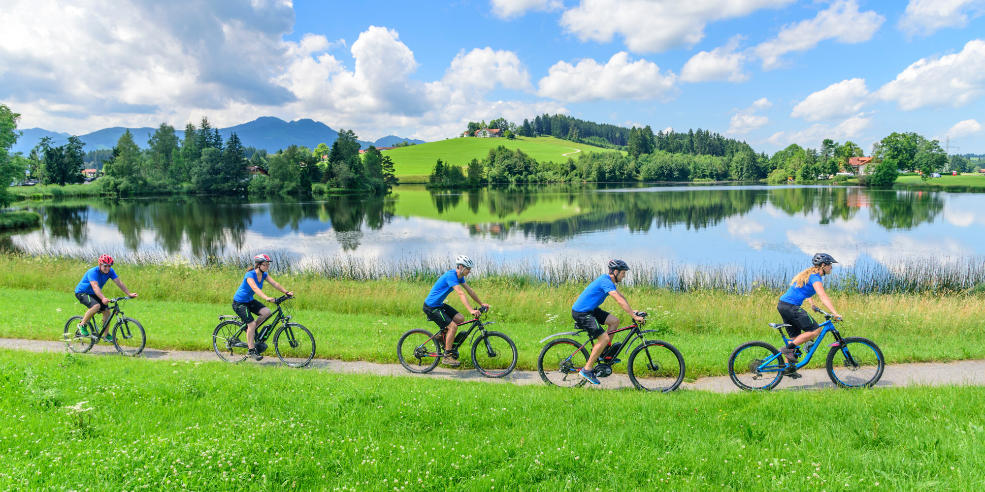 Fünf Radfahrer in blauen T-Shirts fahren an einem See entlang, umgeben von grüner Landschaft.