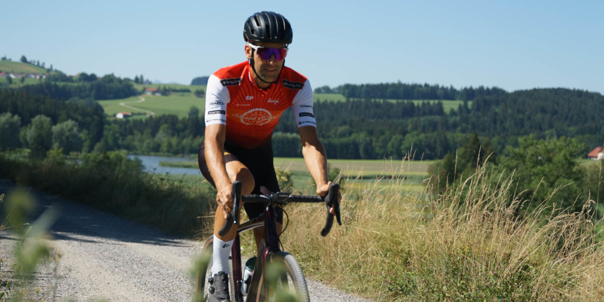 Radfahrer in rot-weißer Kleidung fährt auf einem Schotterweg durch eine Landschaft.