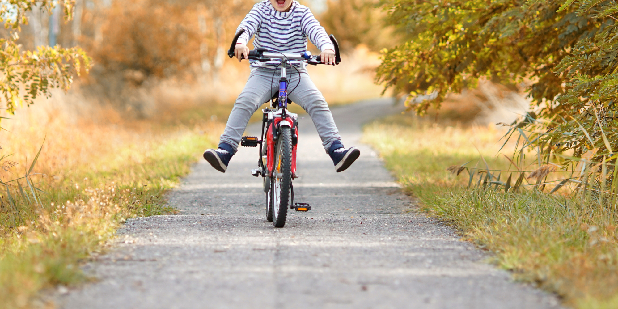 Junge fährt glücklich mit dem Fahrrad auf einem Weg, umgeben von herbstlichen Bäumen.