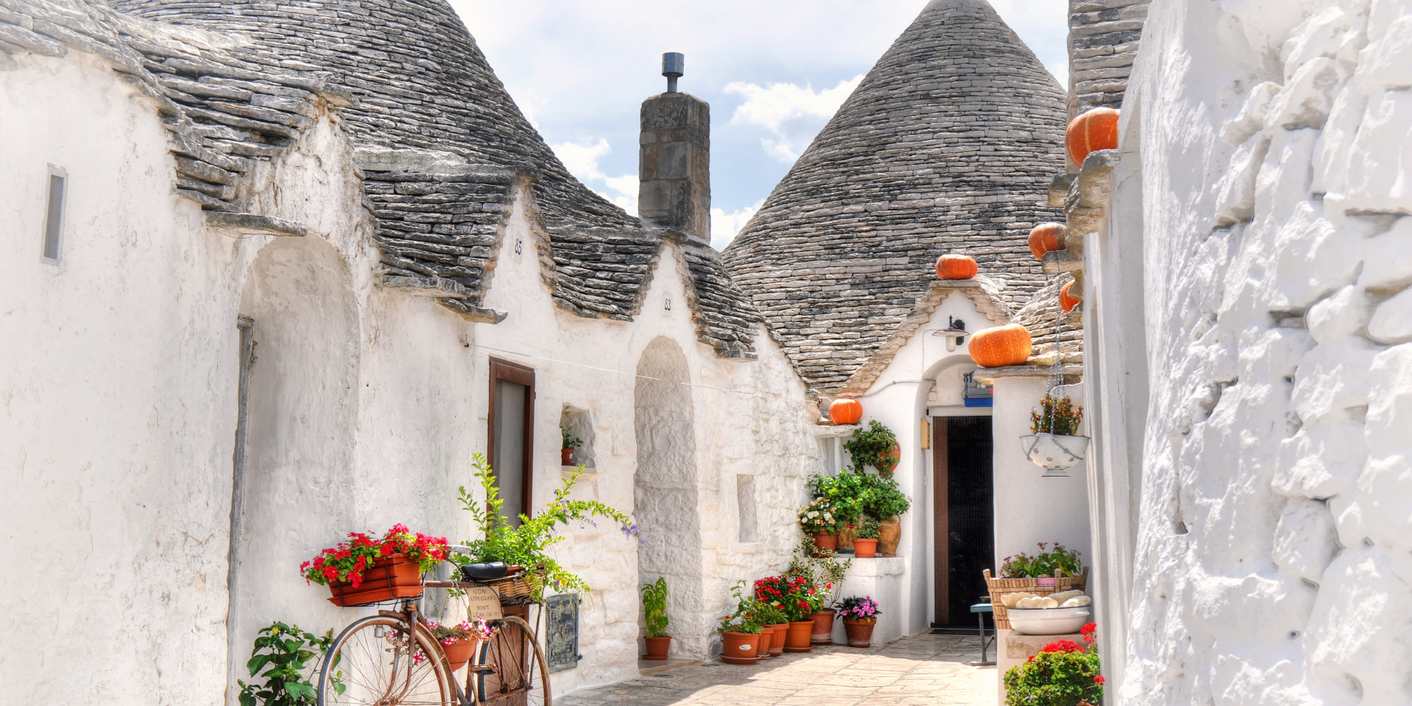 Enggasse mit weißen Trulli-Häusern und bunten Blumen in Töpfen.