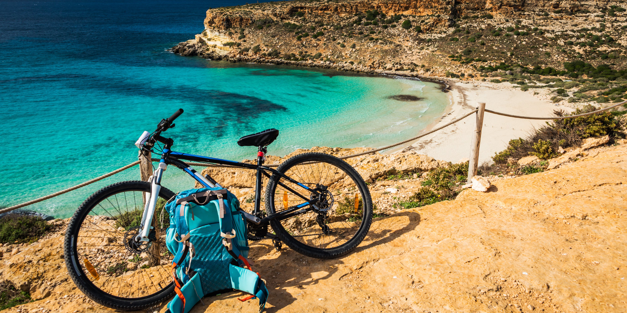 Fahrrad und Rucksack an einem felsigen Küstenweg mit klarem, türkisem Wasser.