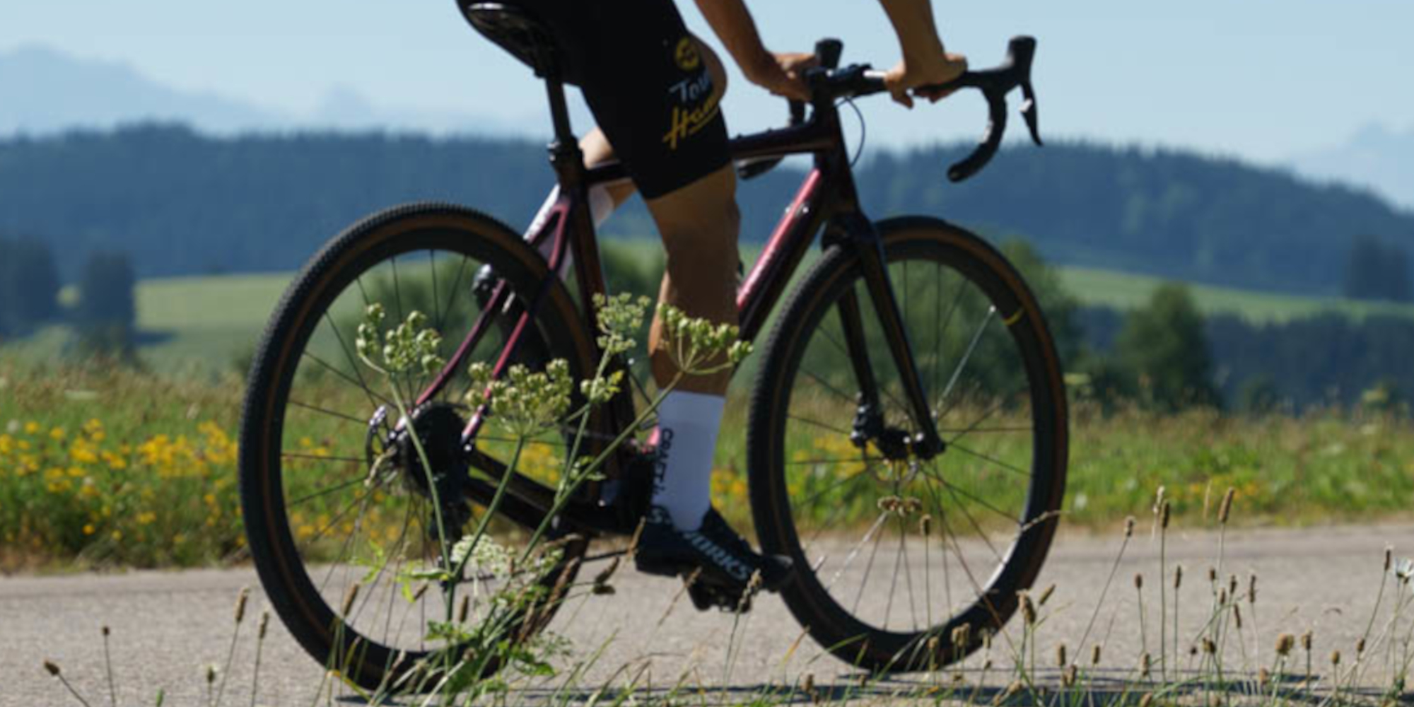Ein Rennradfahrer auf einer Landstraße, umgeben von grünem Gras und Bergen im Hintergrund.