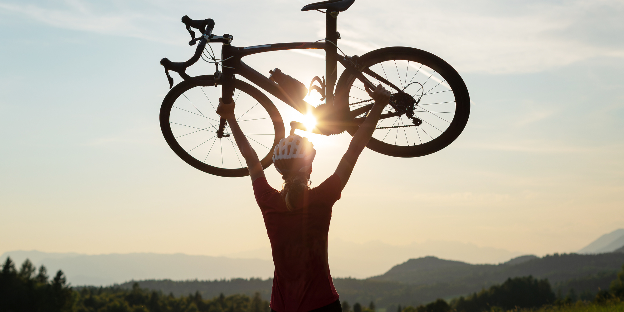 Person hebt ein Fahrrad gegen den Sonnenuntergang in einer bergigen Landschaft.