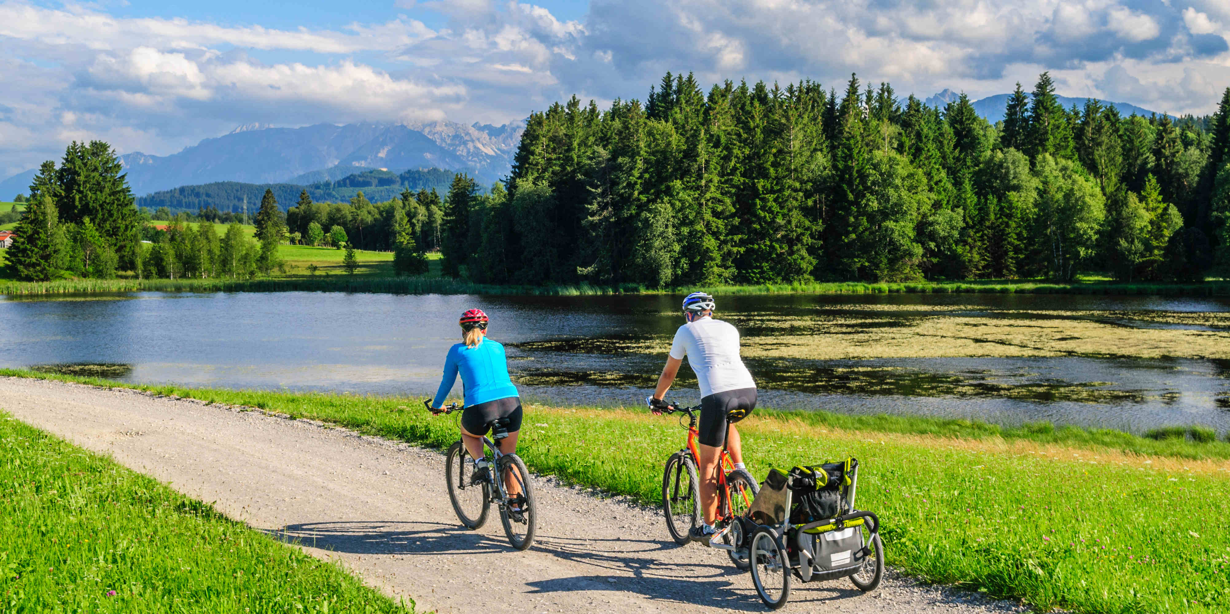 Zwei Radfahrer fahren entlang eines Ufers mit Bäumen und Bergen im Hintergrund.