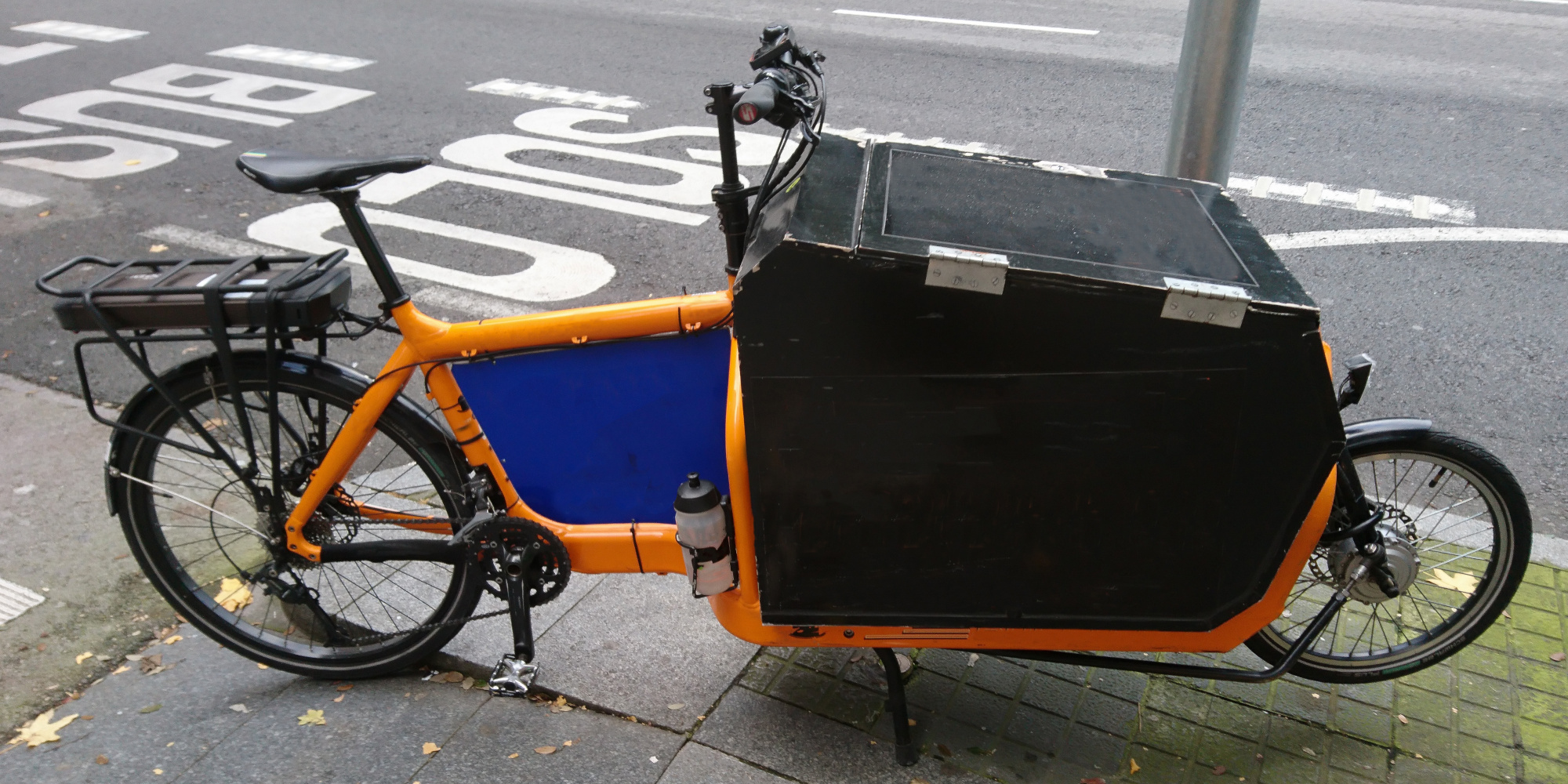 E-Lastenrad Orangenes E-Lastenrad steht an einem Pfosten auf dem Bordstein