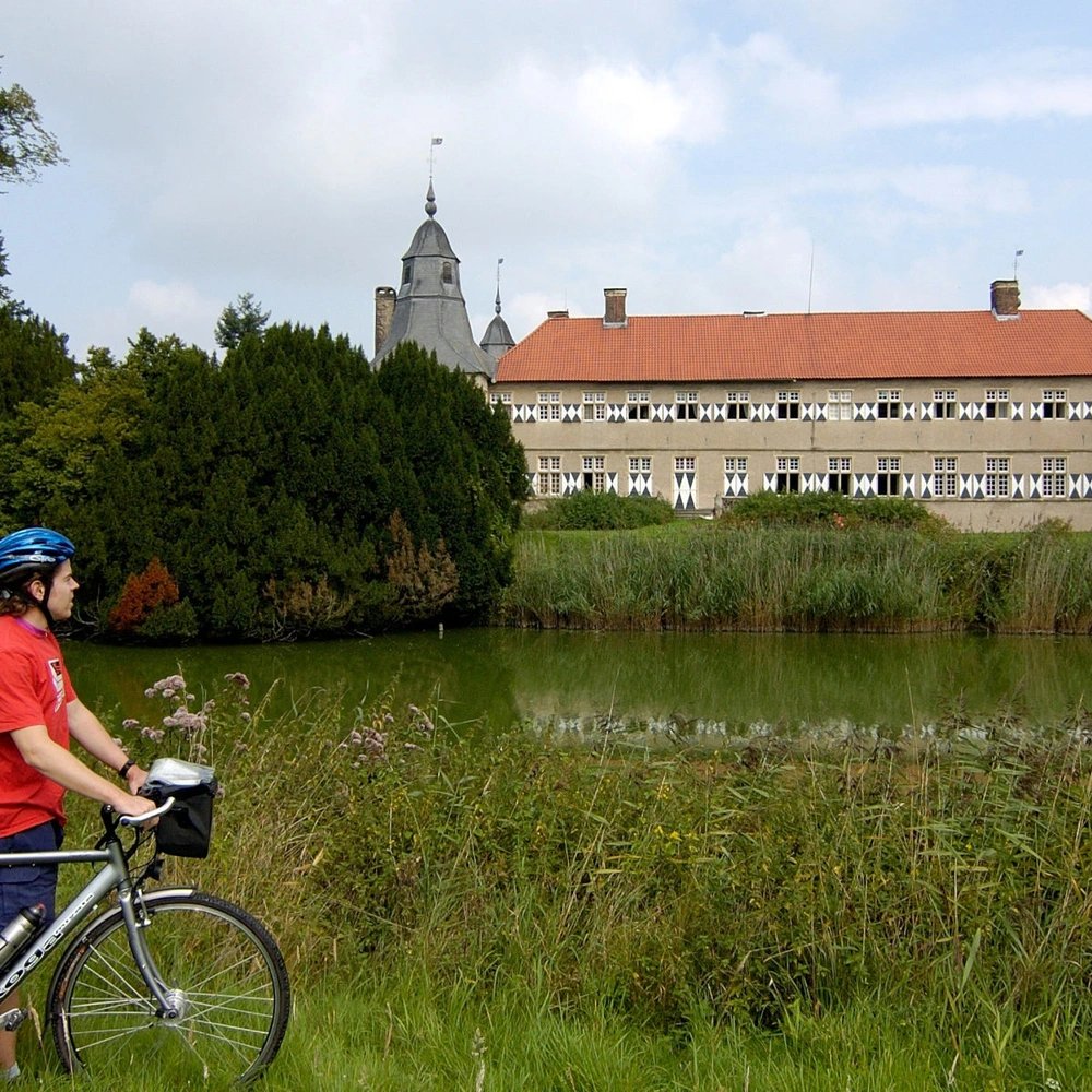 Radfahrer in rot vor einem historischen Gebäude an einem ruhigen Gewässer.