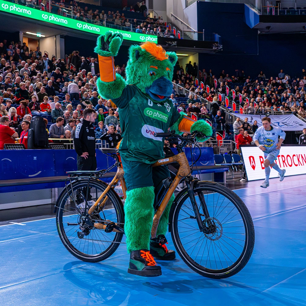 Das Maskottchen der Recken winkt auf einem Fahrrad vor einer jubelnden Menge in der Halle.