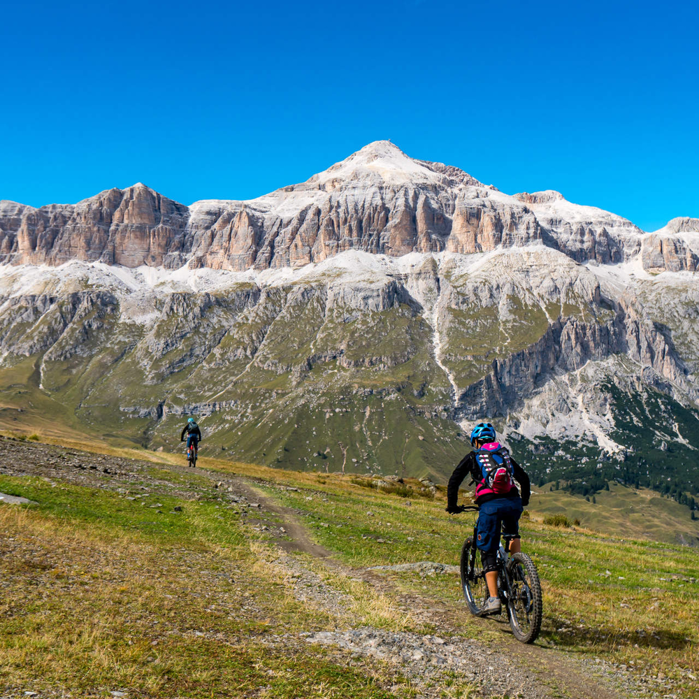 E-Bike Touren in den Dolomiten - schöne Aussicht, vor allem im Sommer Mountainbiker fahren über eine Wiese in Richtung des Sellastocks der Dolomiten
