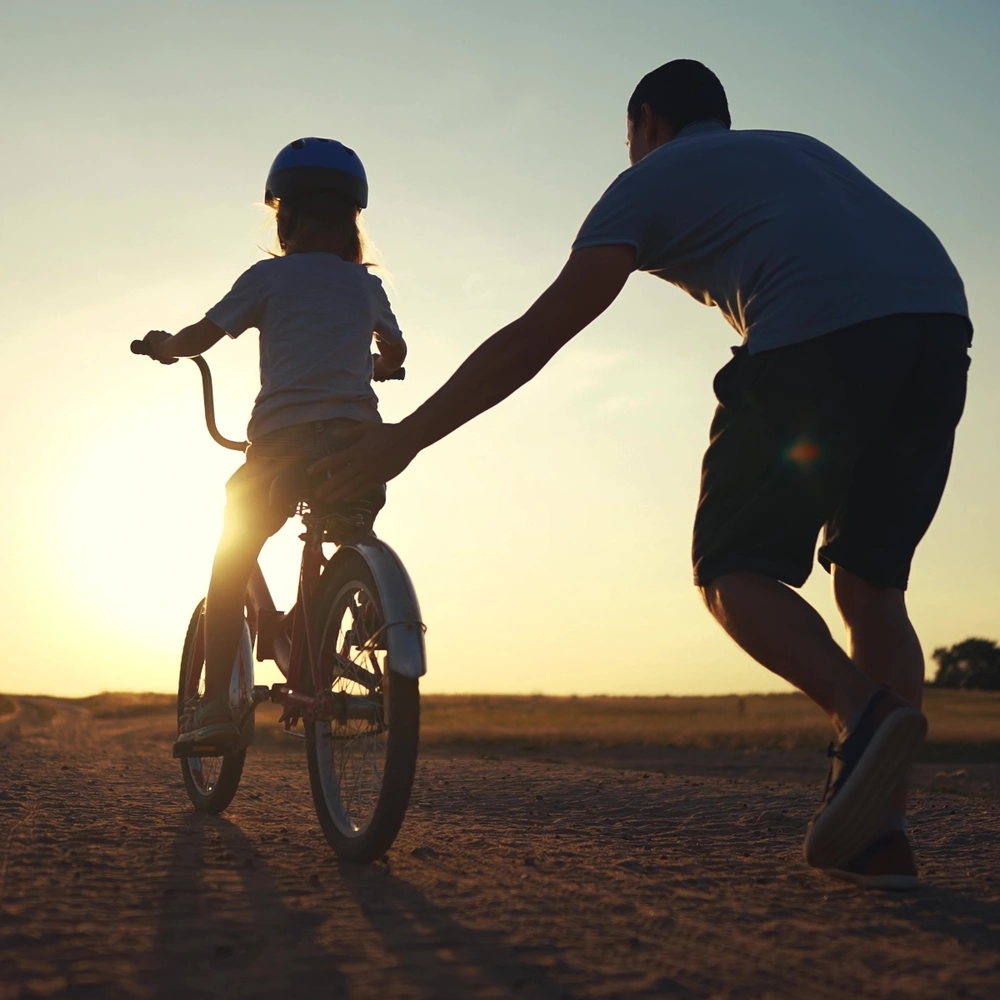Ein Erwachsener hilft einem Kind beim Fahrradfahren, mit Sonnenuntergang im Hintergrund.