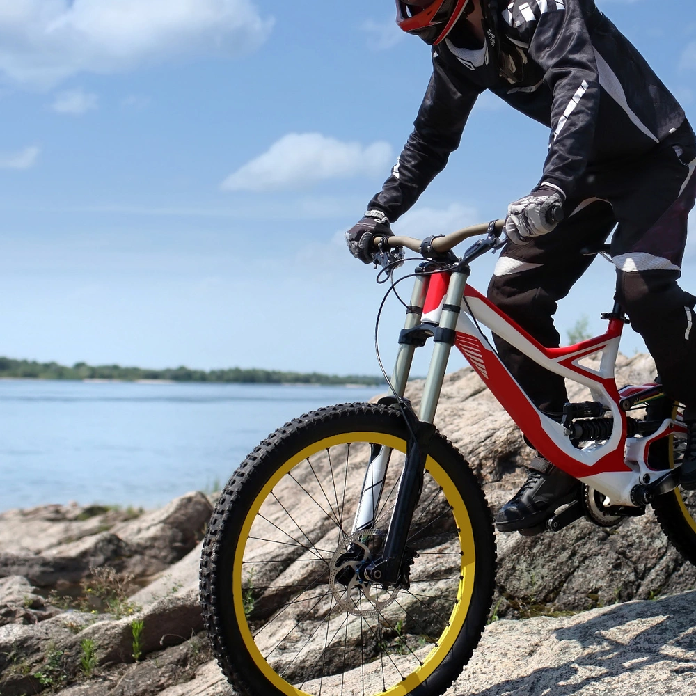 Mountainbiker auf Felsen mit gelben Reifen, im Hintergrund ein Gewässer und blauer Himmel.