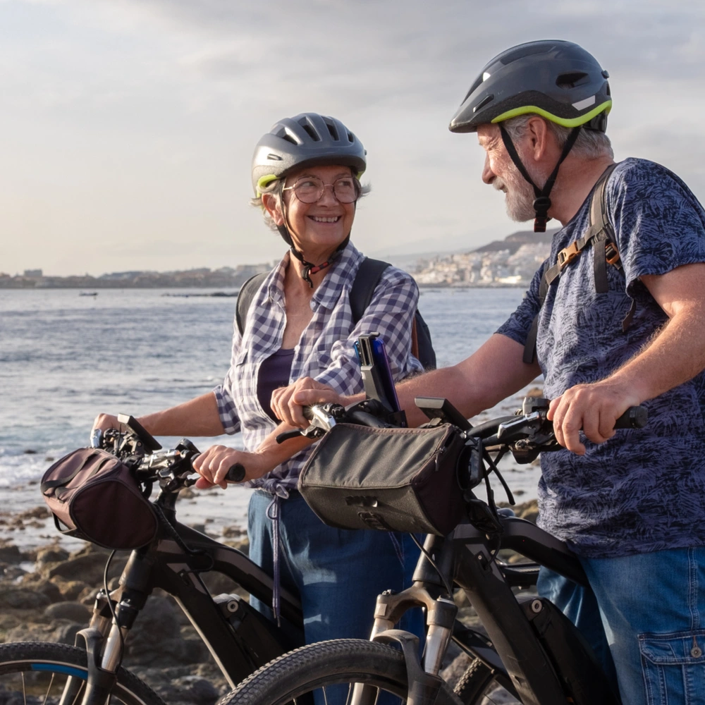 Älteres Paar mit E-Bikes an der Küste, lächelnd und im Gespräch.