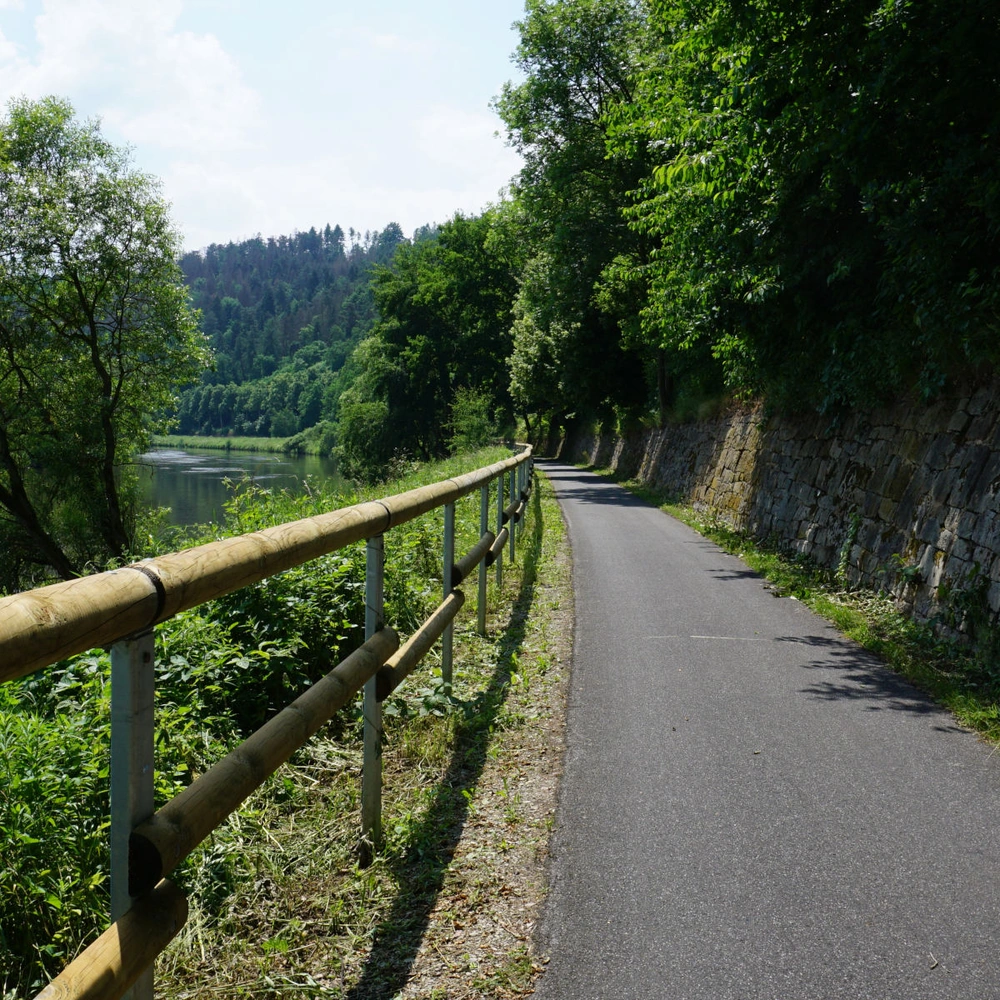 Radweg entlang eines Flusses, flankiert von Bäumen und einer Steinmauer.
