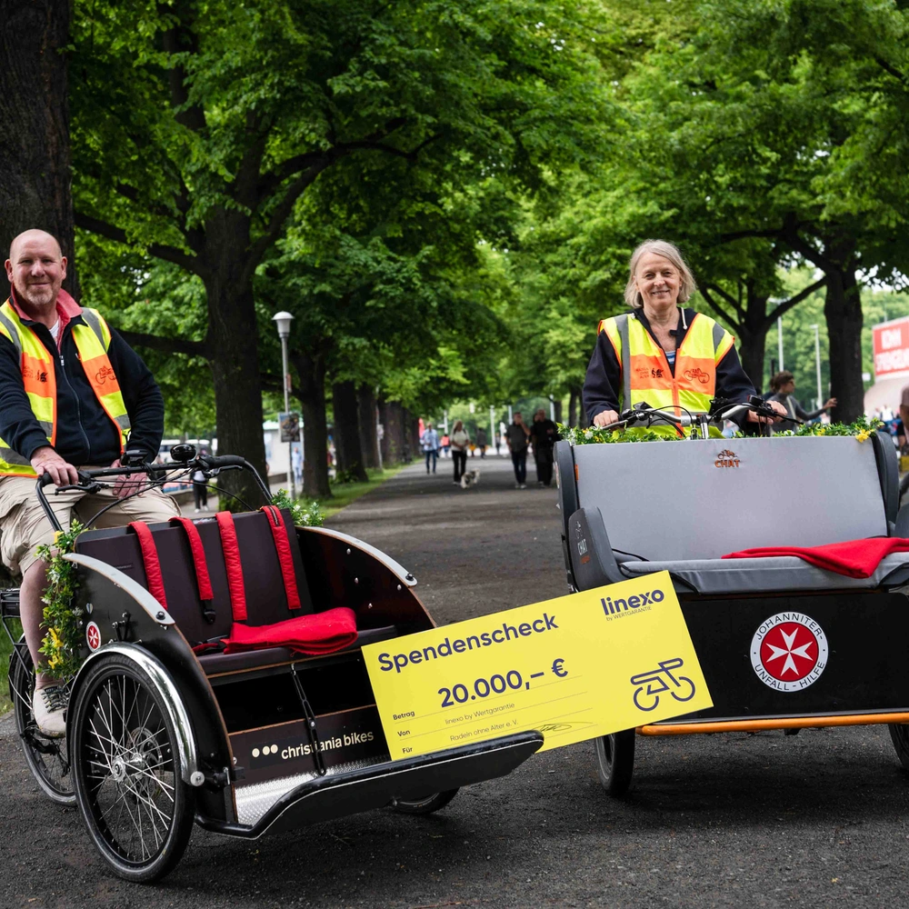 Zwei Personen in Warnwesten stehen neben einer Fahrrad-Rikschawagen auf einem grünen Weg.