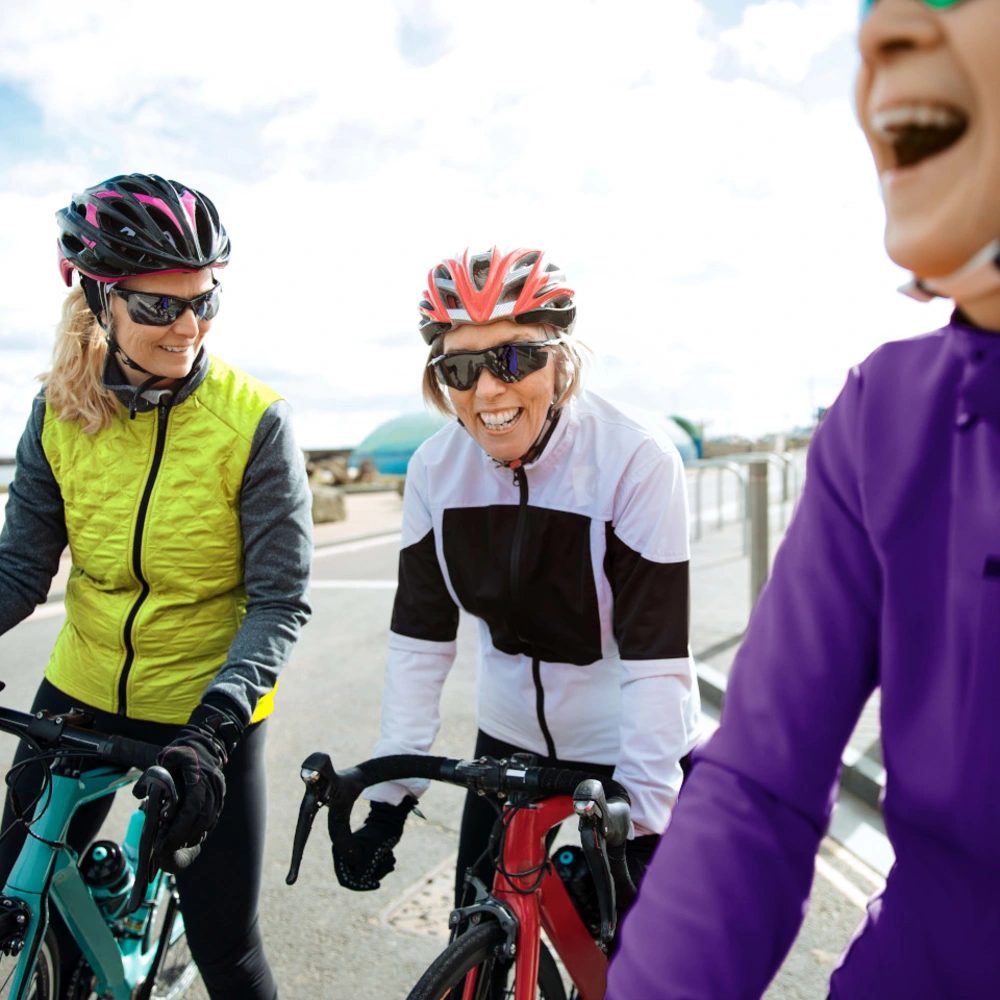 Drei Radfahrerinnen in bunten Sportoutfits lachen auf einer Straße bei bewölktem Himmel