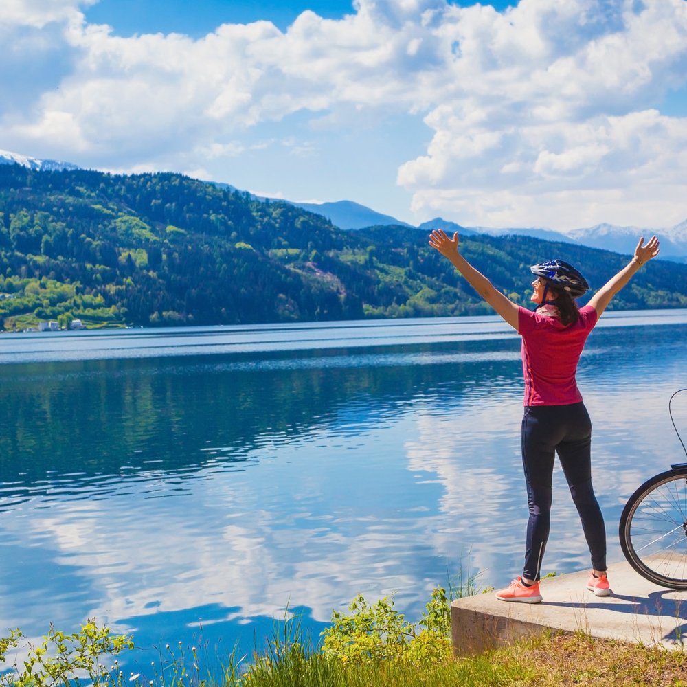 Entdecke die Top 10 Radrouten in Österreich Frau steht vor einem Österreicher See neben einem Fahrrad und hebt ihre Arme freudig nach oben
