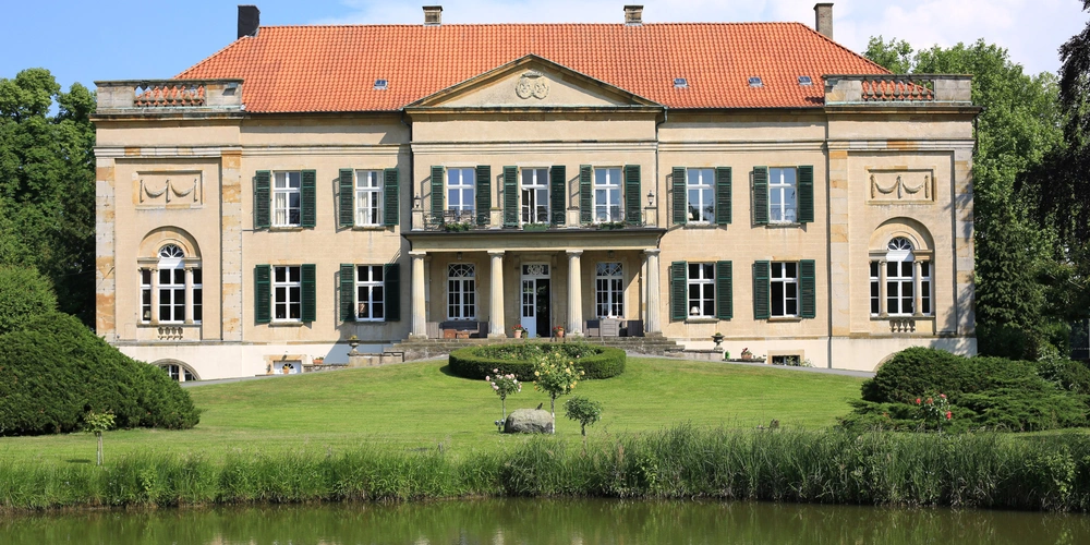 Historisches Gebäude mit grünem Garten und Wasseransicht im Vordergrund.