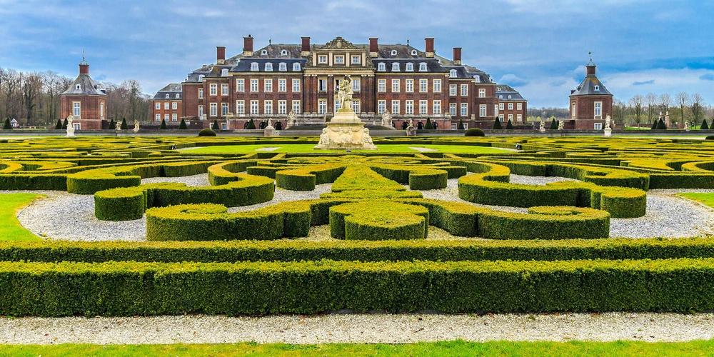 Schloss mit barockem Garten, symmetrische Hecken und Grüntöne unter bewölktem Himmel.