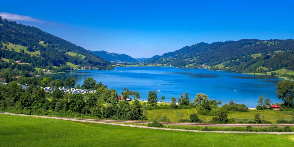 Panoramablick auf einen ruhigen See, umgeben von grünen Hügeln und blauen Himmel.