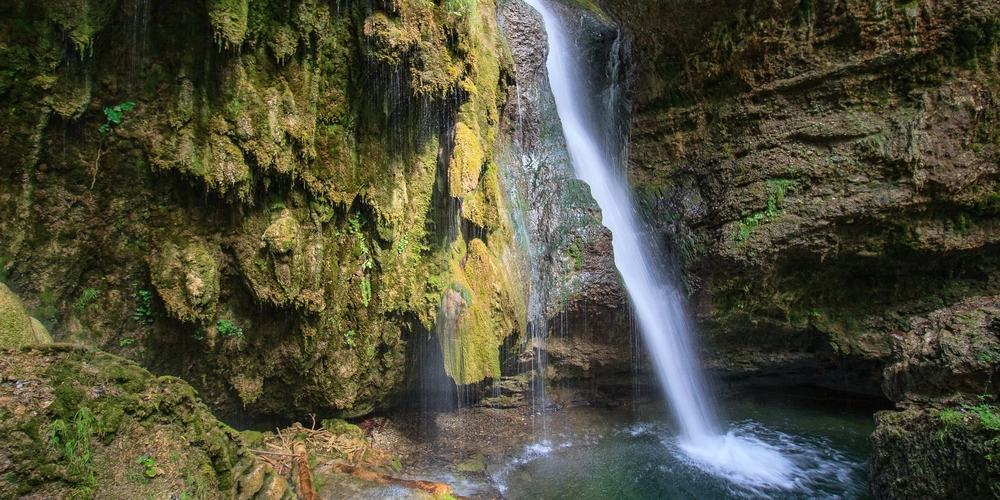 Wasserfall fließt in einen klaren Teich, umgeben von moosbedeckten Felsen.