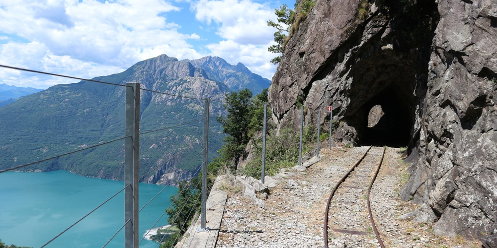 Blick auf einen schmalen Bahnweg mit Tunnel, umgeben von Bergen und Wasser.