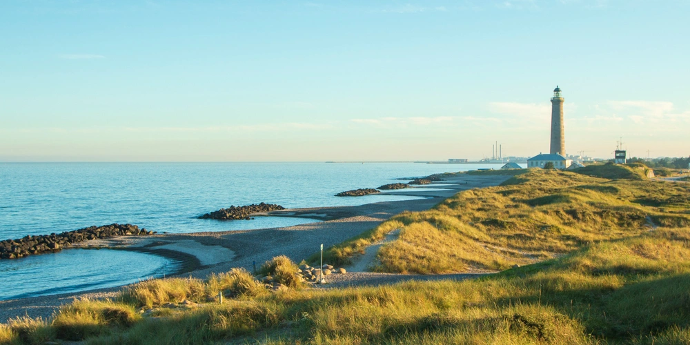 Küstenlandschaft mit Leuchtturm, sanften Dünen und ruhigem Wasser.