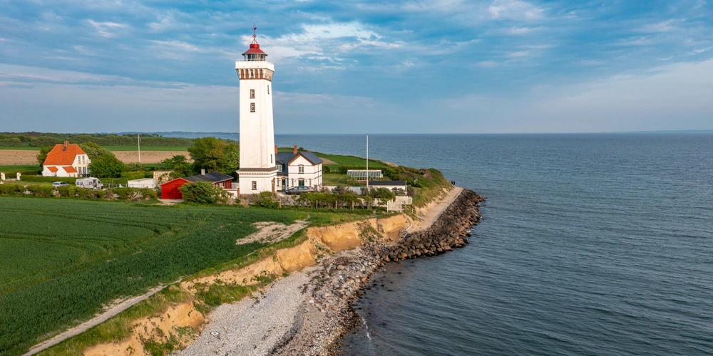 Leuchtturm an der Küste mit grünem Ufer und blauem Himmel.