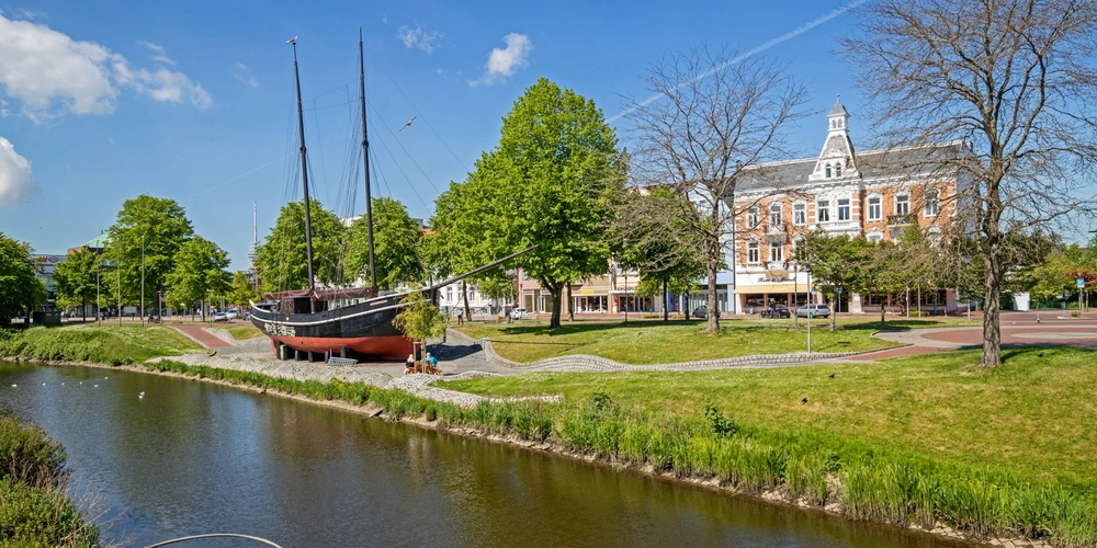 Park mit einem Schiff, Bäumen und einem Gebäude im Hintergrund an einem ruhigen Wasserlauf.