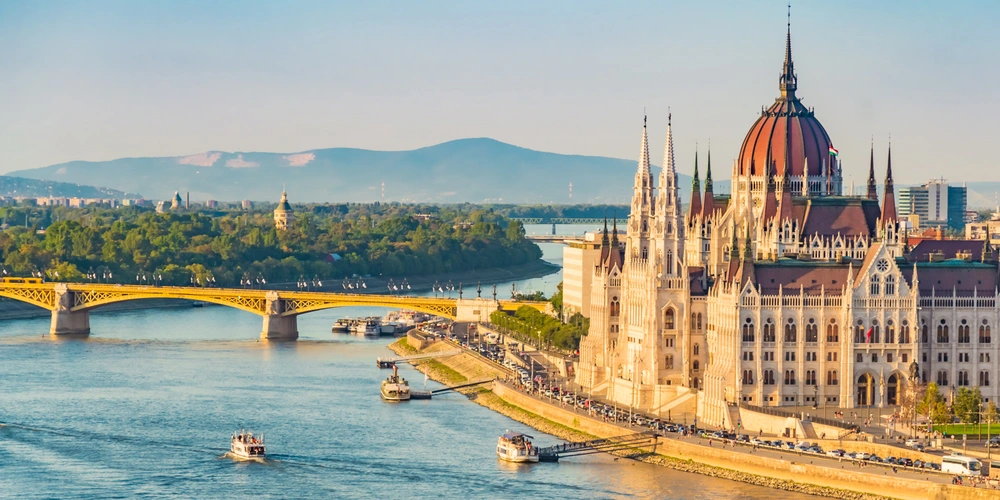 Blick auf das ungarische Parlamentsgebäude am Donauufer mit Brücke im Hintergrund.