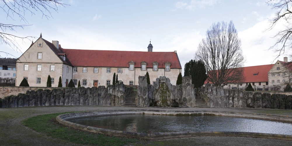 Historisches Gebäude mit rotem Dach und Brunnen in der Landschaft.