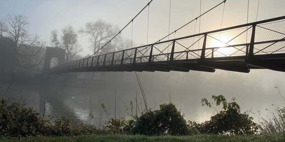 Hängende Brücke über einem ruhigen, nebligen Gewässer bei Sonnenaufgang.