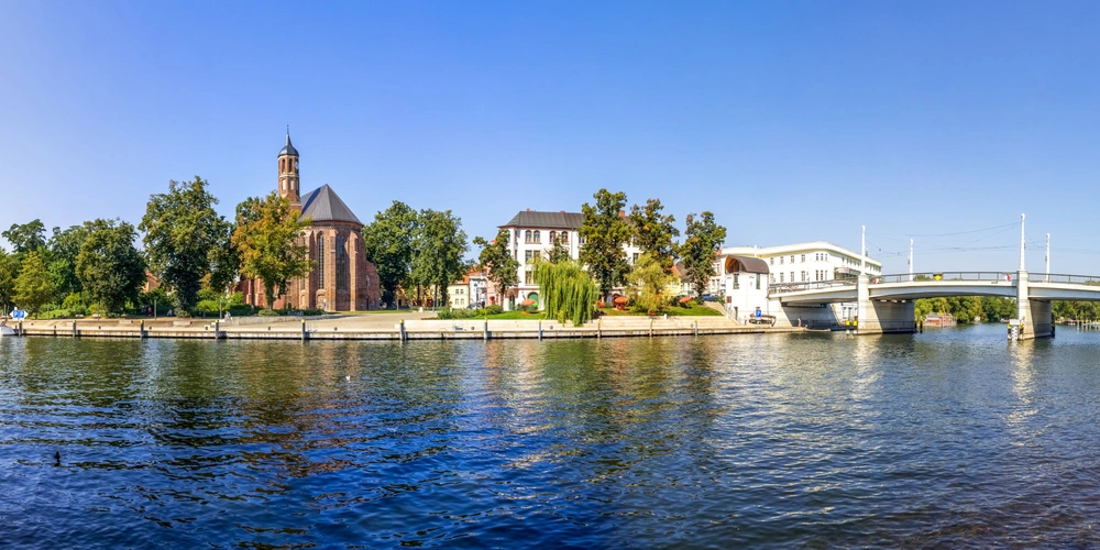 Uferansicht mit einer Kirche, Bäumen und einer modernen Brücke bei klarem Himmel.