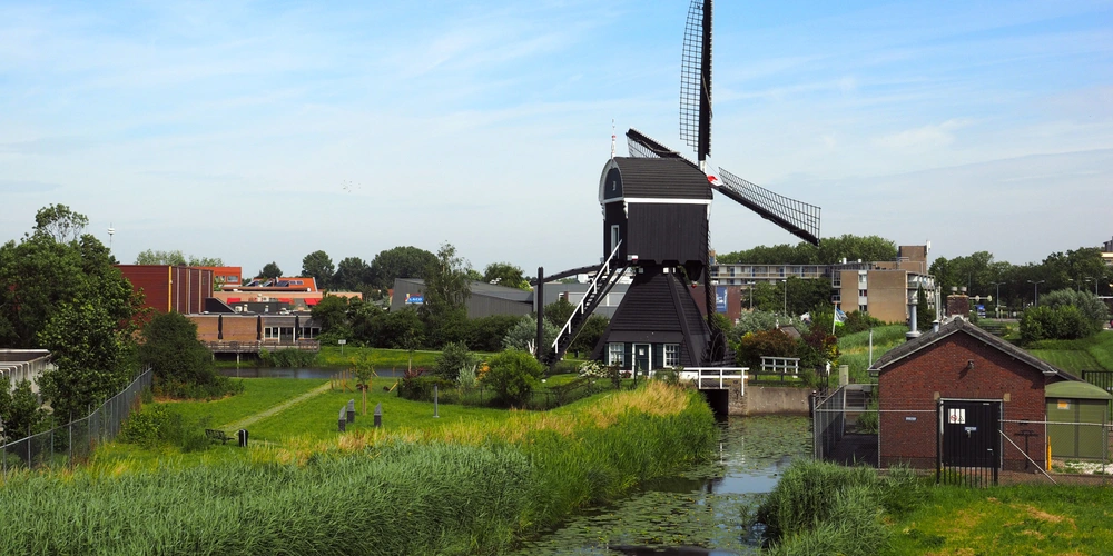 Schwarze Windmühle neben einem Fluss mit grüner Landschaft und Häusern im Hintergrund.