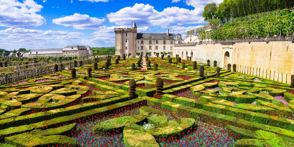 Historische Gartenlandschaft mit geometrischen Formen und bunten Blumen vor einem Schloss.