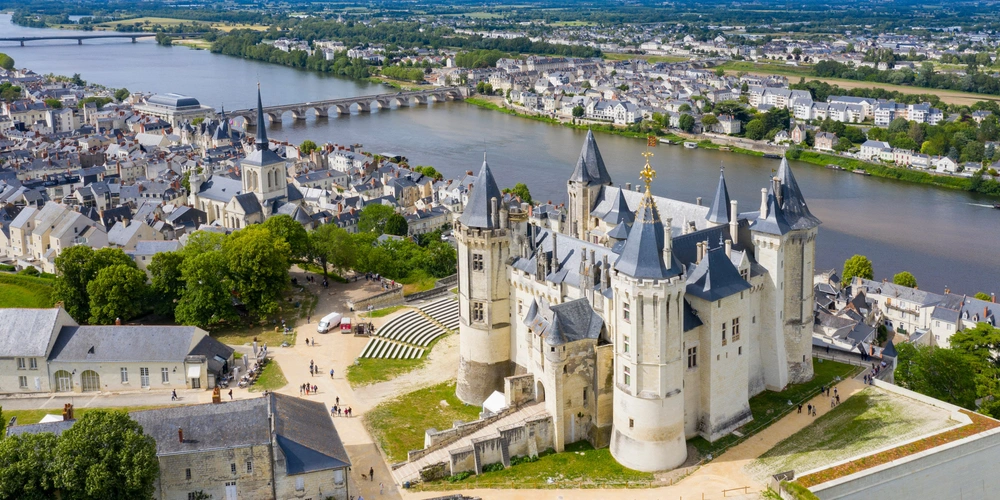 Schloss mit Türmen und Zinnen, umgeben von grünen Bäumen und einem Fluss im Hintergrund.