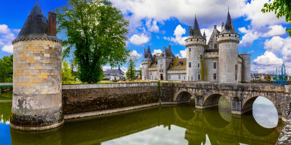 Schloss mit Türmen, Brücke und Teich, umgeben von grüner Landschaft und blauem Himmel.