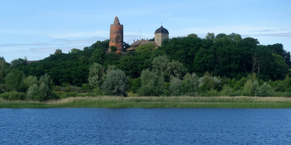 Der Rote Turm Pouch  Blick auf eine Burg auf einem Hügel, umgeben von Bäumen und einem ruhigen See.