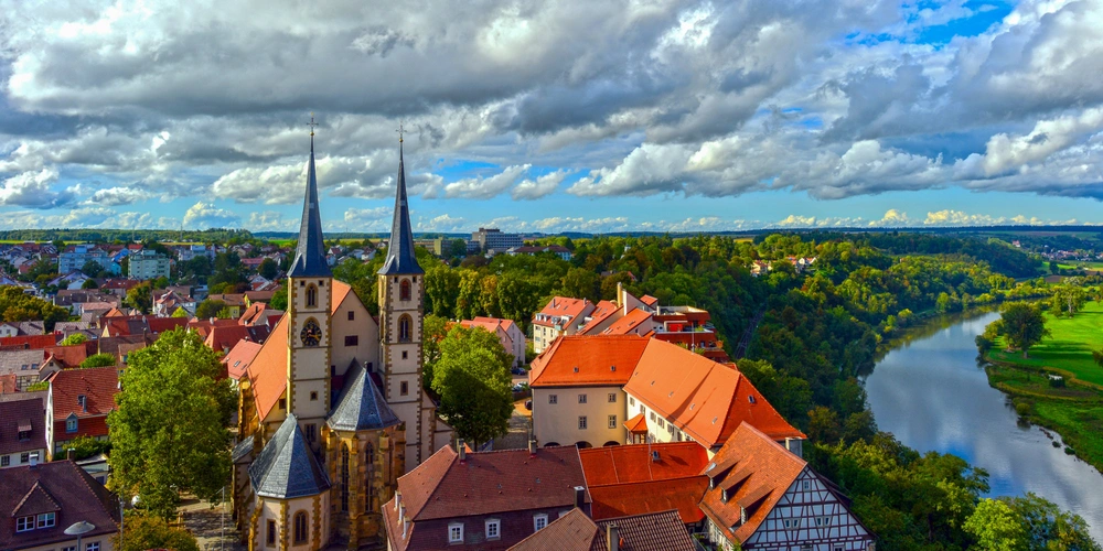 Altstadt von Bad Wimpfen Blick über eine Stadt mit Kirchtürmen, fließendem Fluss und grünen Hügeln unter bewölktem Himmel.