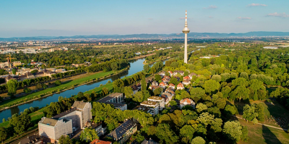 Mannheim Luftaufnahme einer Stadt mit Fluss, Bäumen und einem Fernsehturm im Hintergrund.