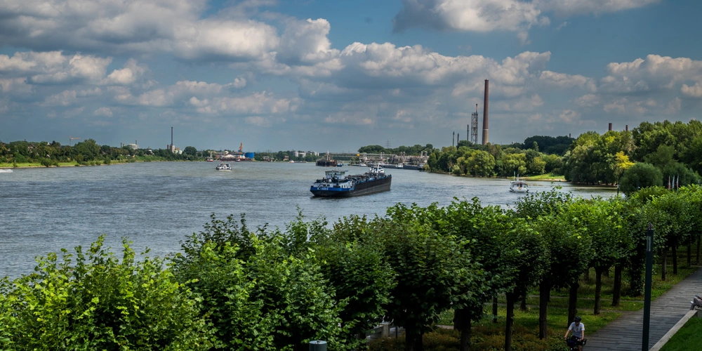 Flusslandschaft mit einem Schiff, Bäumen und einer industriellen Kulisse im Hintergrund.