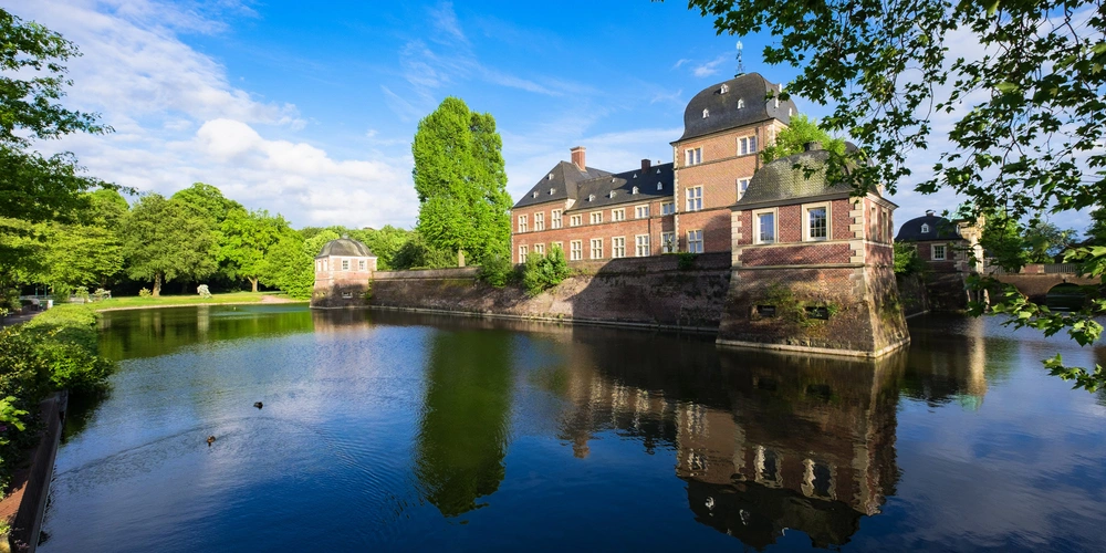 Schloss mit Wasserreflexion in Ahaus, umgeben von Bäumen und blauem Himmel.