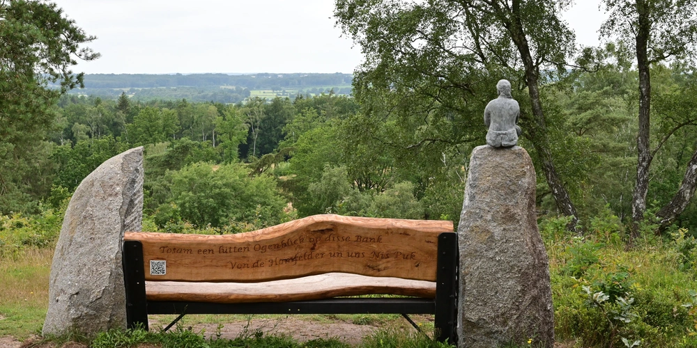 Holzbank zwischen zwei Steinen mit einer Skulptur im Hintergrund und natürlicher Landschaft.