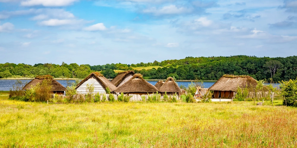 Bauernhäuser mit Strohdächern in einer grünen Wiese am Wasser, blauer Himmel im Hintergrund.