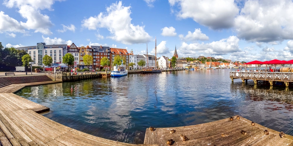 Flensburg Hafen Hafenansicht mit Booten, Holzsteg und bunten Gebäuden unter blauem Himmel.