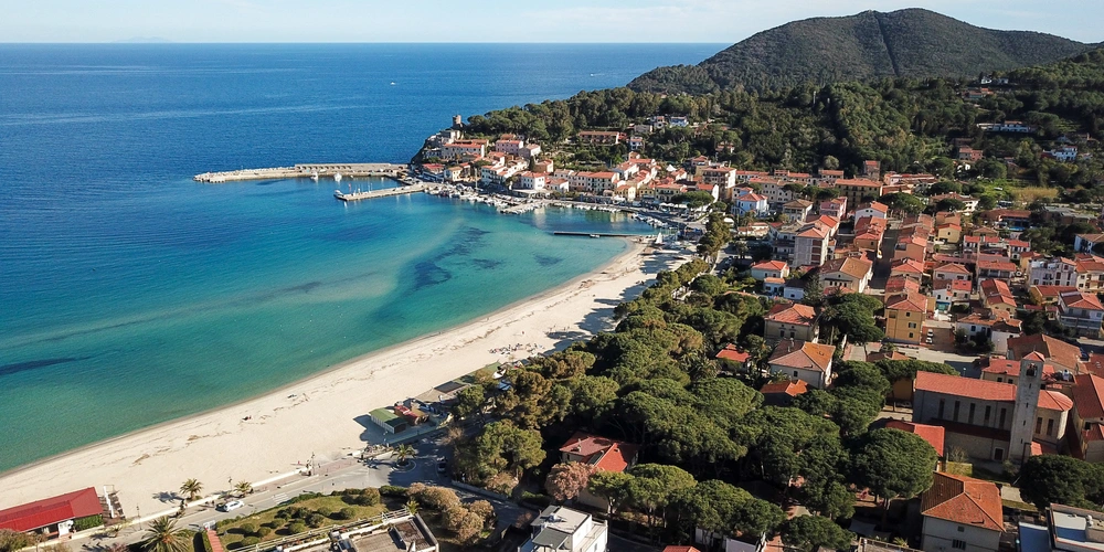 Luftaufnahme einer Küstenstadt mit Strand, klarem Wasser und grünen Hügeln im Hintergrund.